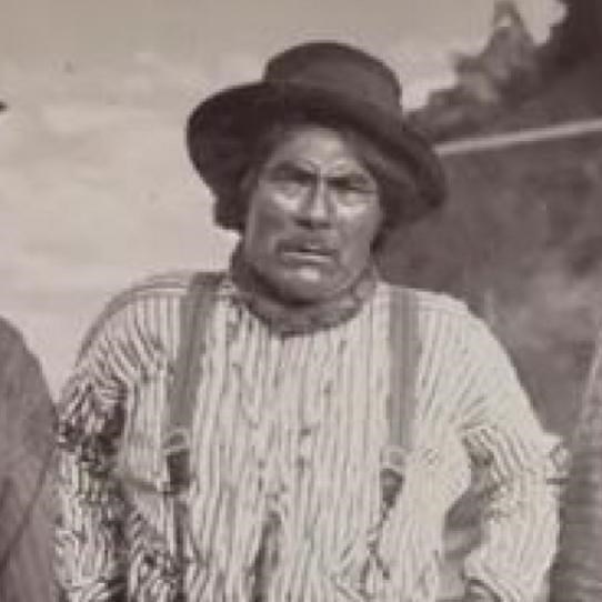 In a historic scene, three men stand on a bank beside a river. Each wears a hat and button up shirt.